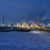 A bridge over a snow-covered river lit up at night, seen from afar.