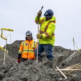Two construction workers standing on top of a mound of soil. One construction worker is hammering a wooden stake into the mound.