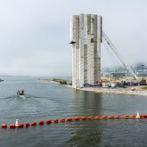 Panoramic view of a large urban construction site for a vertical lift bridge over a river, with a boat passing by and a foggy city skyline in the background.