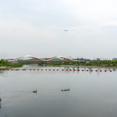 View of a serene river with a red bridge in the background, under a cloudy sky. An airplane is visible in the sky, and a row of ducks swims in the water.
