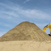A pile of soil against a blue sky. An excavator is half hidden behind the pile.