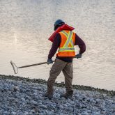 A worker in high-visibility construction clothing holding a rake next to a river.