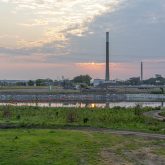 Sunset over a tranquil river with reflections of the sky and clouds, surrounded by lush greenery. In the background, there are power lines and a smokestack.