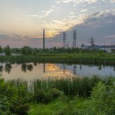 Sunset over a calm river with reflections of the sky and surrounding foliage, featuring a distant industrial tower and overhead power lines, with a train passing on a nearby elevated track.