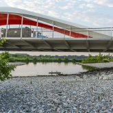 View of a modern pedestrian bridge over a river, featuring a sleek white design with a vibrant red railing, set against a backdrop of a calm river and greenery.