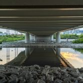 View from under a bridge with the reflection of the underside and pillars in a calm river, rocky foreground, and buildings in the distance at dusk.
