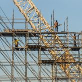 A construction worker stands on top of scaffolding that surrounds a white industrial crane.