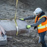 A construction worker pushing a large boulder suspended by a rope.