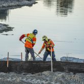 Two construction workers raking by the water.