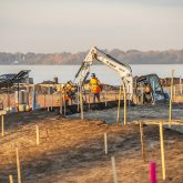 A construction worker framed by a small excavator with the lake in the background.