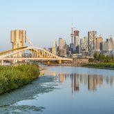 Panoramic view of the Toronto skyline featuring the CN Tower and a modern yellow bridge over a reflective body of water with foreground greenery.