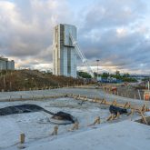 View of a construction site with a large cement plant and silos under a cloudy sky. The foreground features unfinished groundwork supported by wooden stakes.