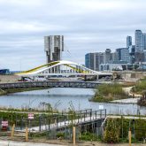 View of a construction site featuring a white and yellow bridge in the foreground, with the CN Tower and downtown Toronto skyline in the background. A small body of water and some greenery are visible in the scene.