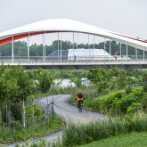 View of a modern bridge with a distinctive red and white arch, crossing over a lush green park where a cyclist and pedestrians are enjoying the path below.