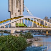 A green train crossing a modern white and yellow arched bridge over a river with city skyscrapers in the background.