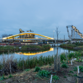 The lights of a bridge reflected in the water of a river. A crane in the background is also lit up.